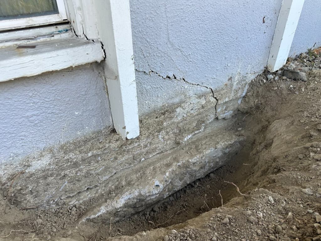 Exterior diagnostic trench excavation along the perimeter foundation footing showing diagonal stucco cracking and foundation exposure
